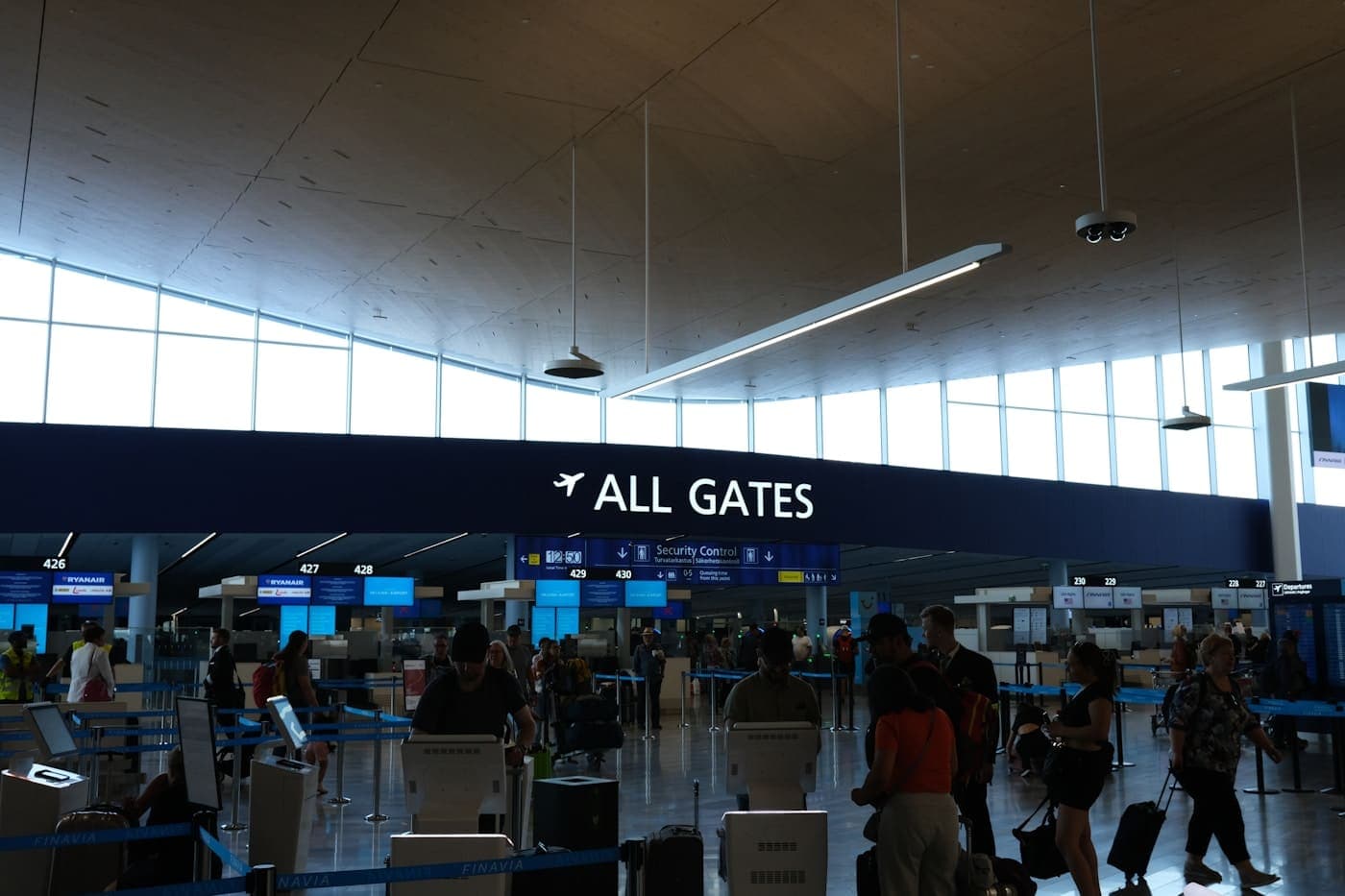 Travellers walking through a bright airport terminal beneath a sign reading All Gates.