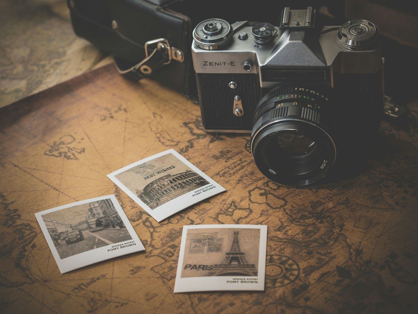A vintage film camera resting on an old paper map, with small printed photographs scattered beside it.