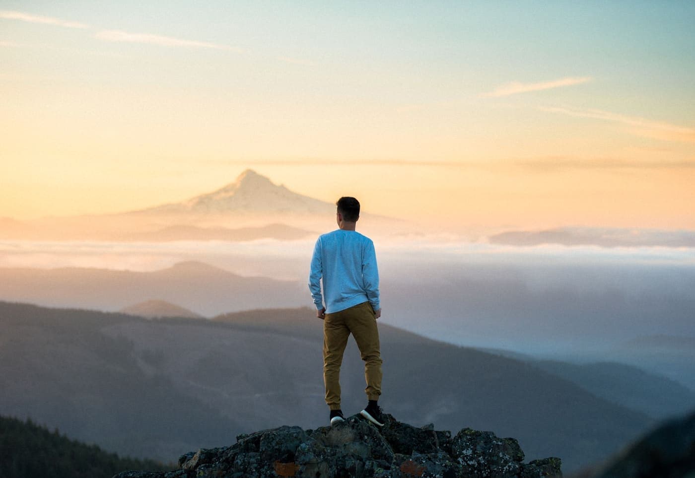 A person standing alone on a rocky outcrop at sunrise, looking toward a distant mountain peak.