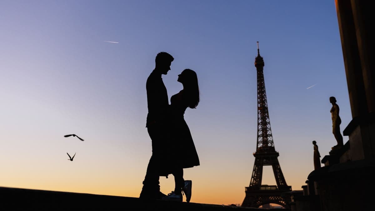 A couple silhouetted against an orange sunset, standing face to face with the Eiffel Tower behind them.