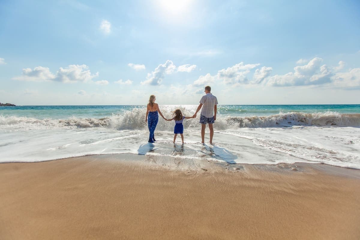 A man, a woman and a small child holding hands at the water’s edge as a wave breaks around them.