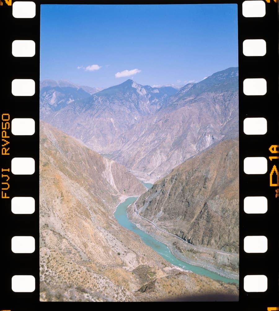 A river winding through a steep, dry mountain valley, framed by the sprocket holes of a 35mm film strip.