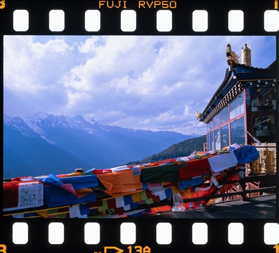 Strings of colourful prayer flags in front of a temple roof and snow-capped peaks, framed by the sprocket holes of a 35mm film strip.