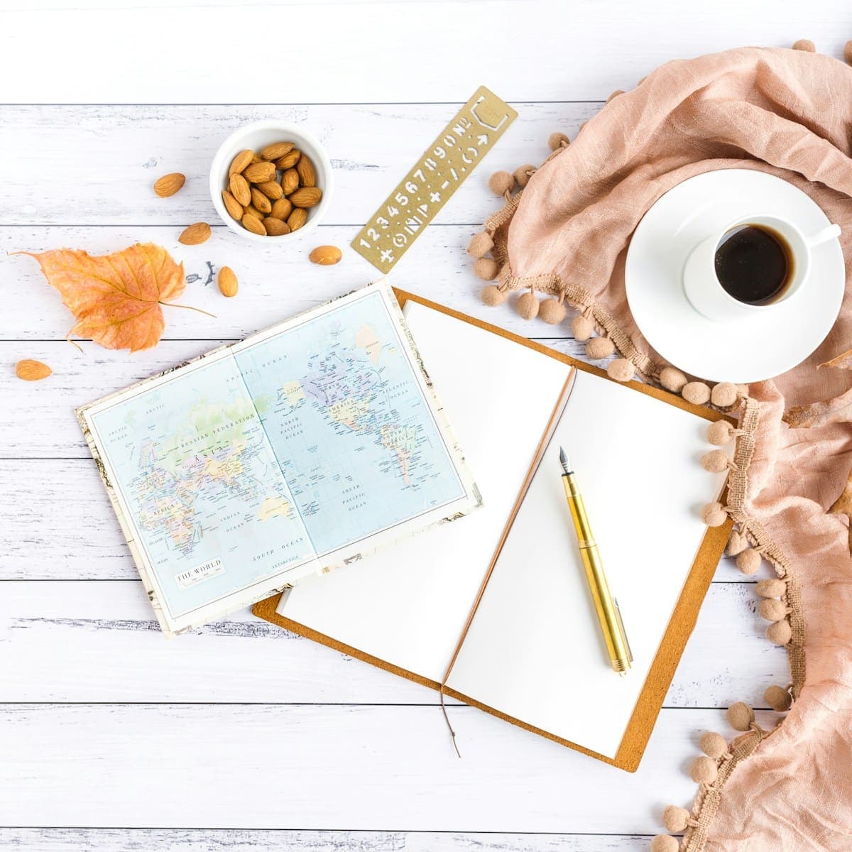An open journal, a folded paper map, a fountain pen and a cup of black coffee laid out on a pale wooden table.